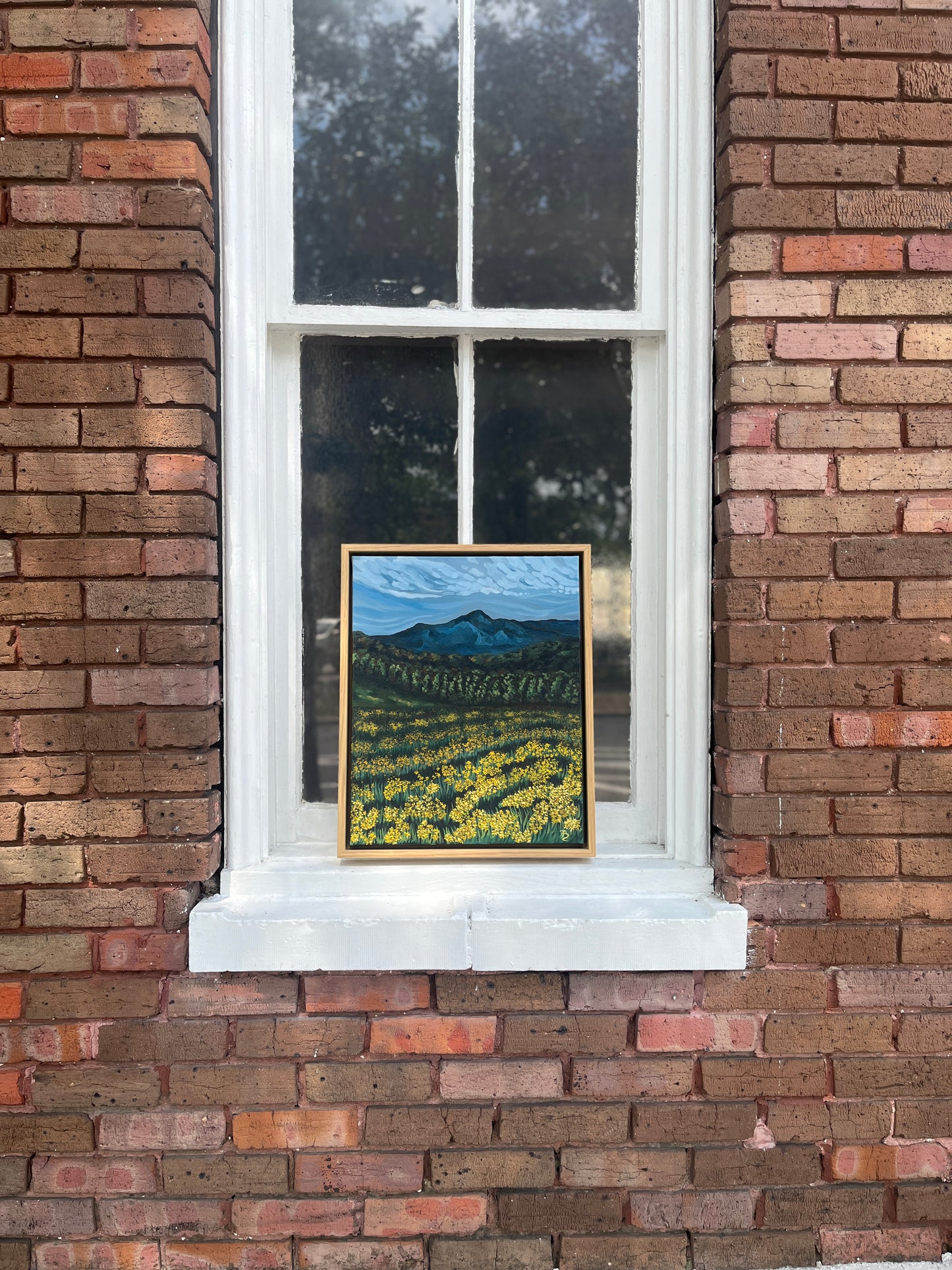 Framed original oil painting of a mountain landscape and meadow of yellow wildflowers. The painting is propped in the windowsill of a historic brick building.