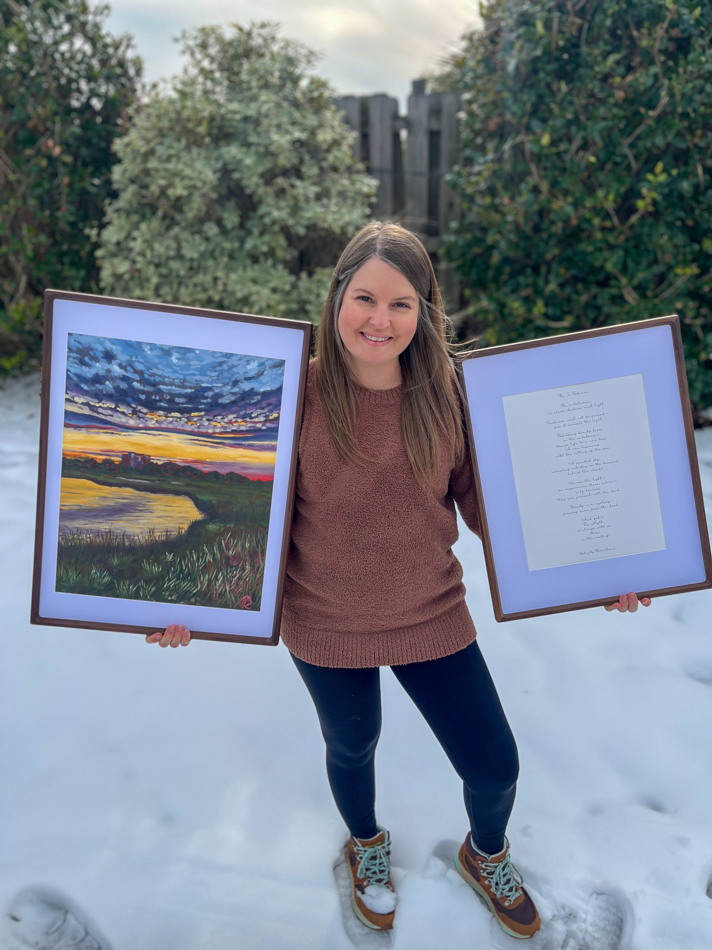 Artist, Blair Owens, holding a framed landscape painting of a sunset over the marsh as well as a framed poem outdoors in the snow
