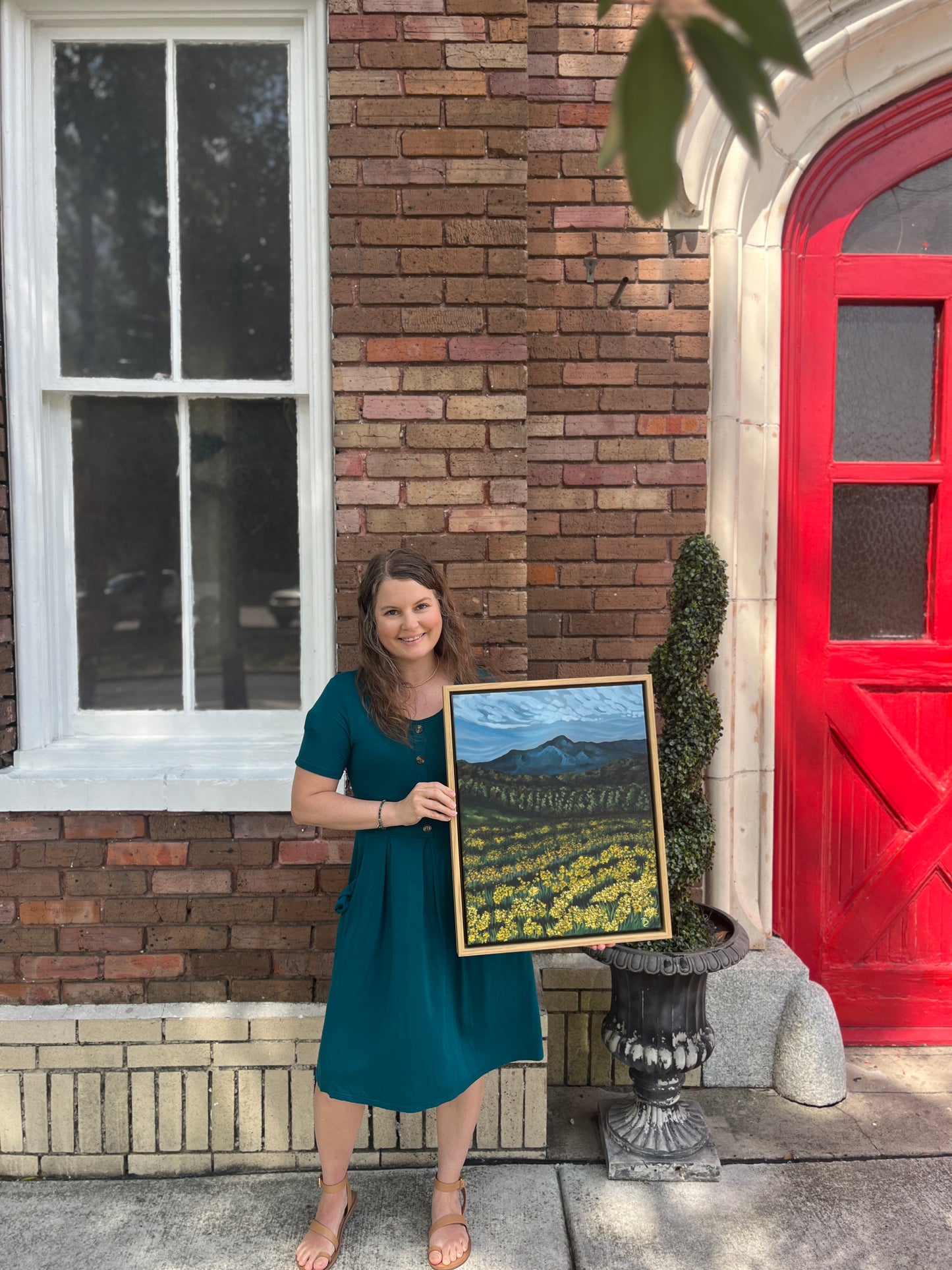 Artist, Blair Owens, holding her framed original oil painting of a mountain landscape while standing outside of a brick building with a red door.