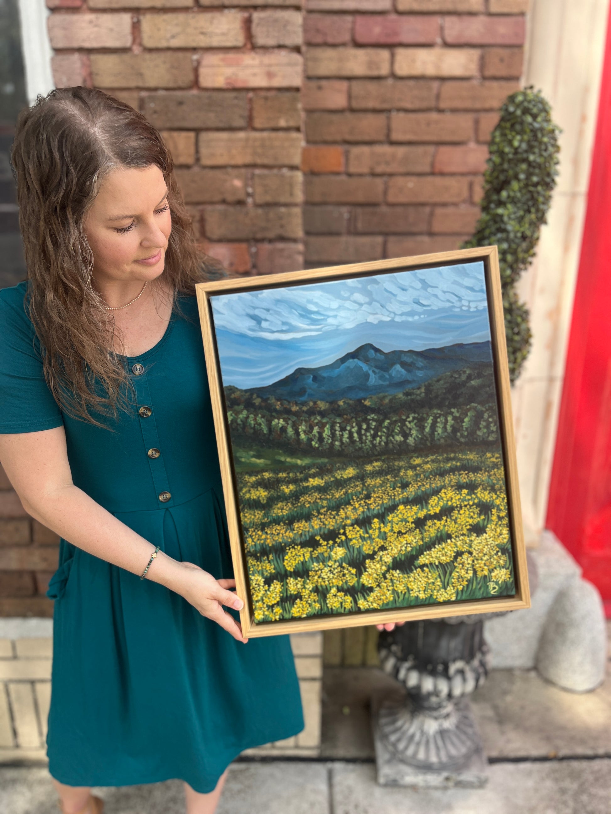 Artist, Blair Owens, holding her framed original oil painting of a mountain landscape while standing outside of a brick building.
