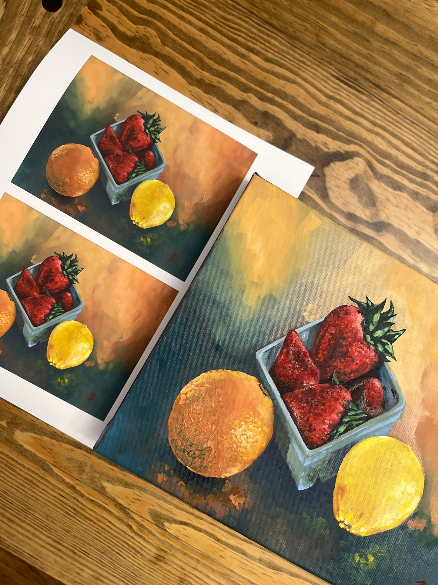 Original still life painting and prints of strawberries, an orange and a lemon on a wooden surface.