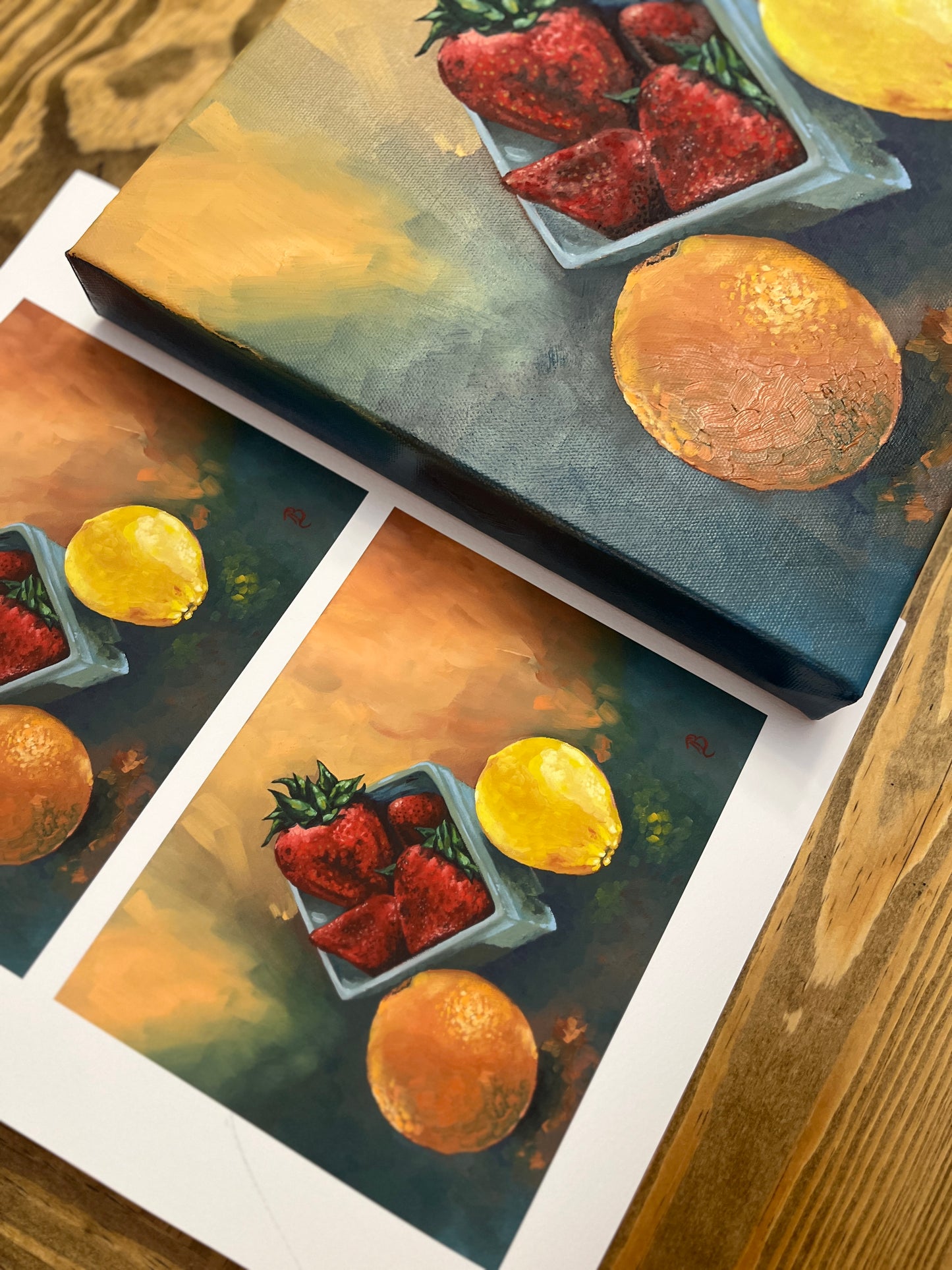 Original still life painting and prints of strawberries, an orange and a lemon on a wooden surface.