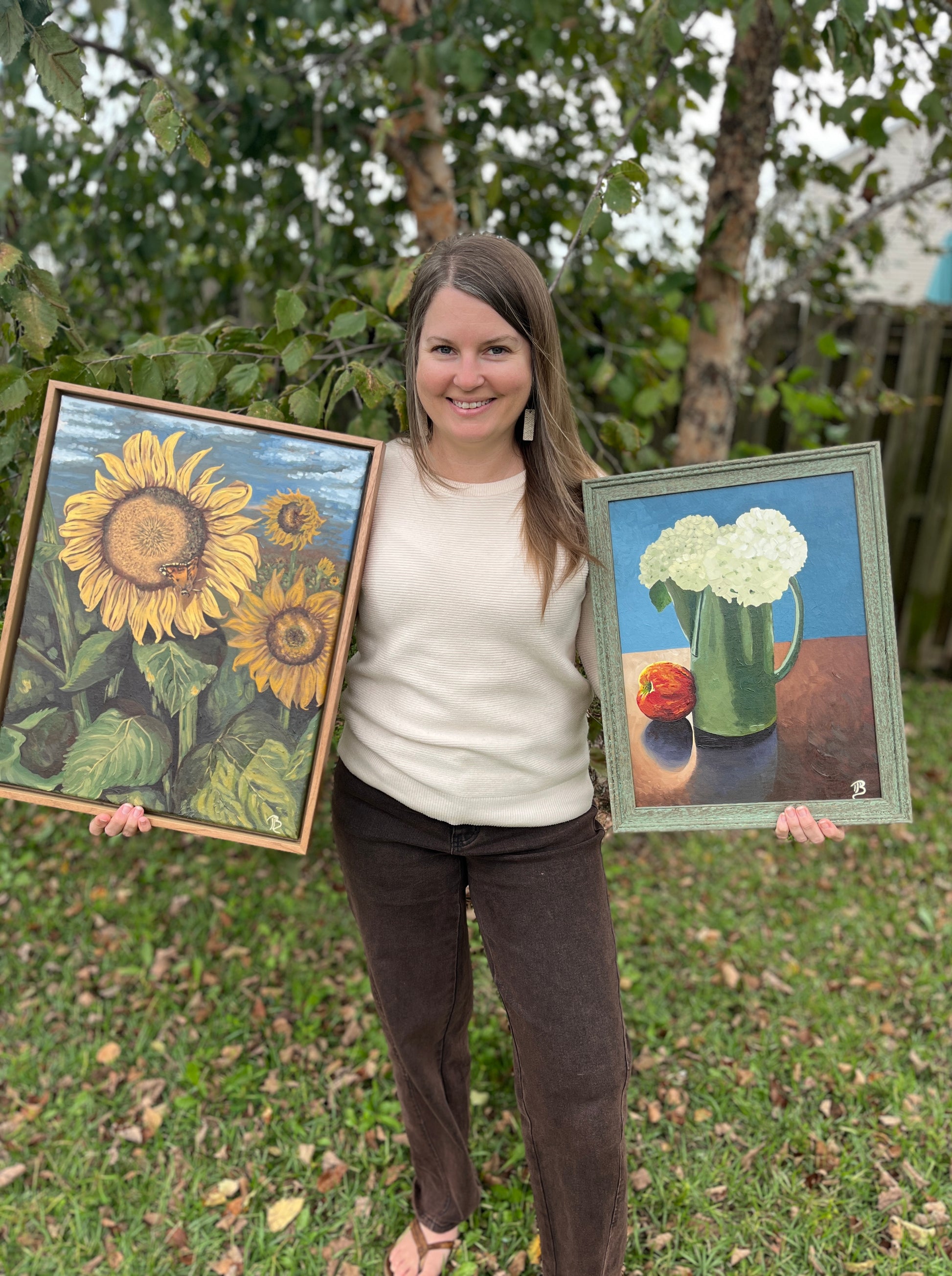 Artist, Blair Owens, holding two framed floral paintings outdoors with greenery in the background
