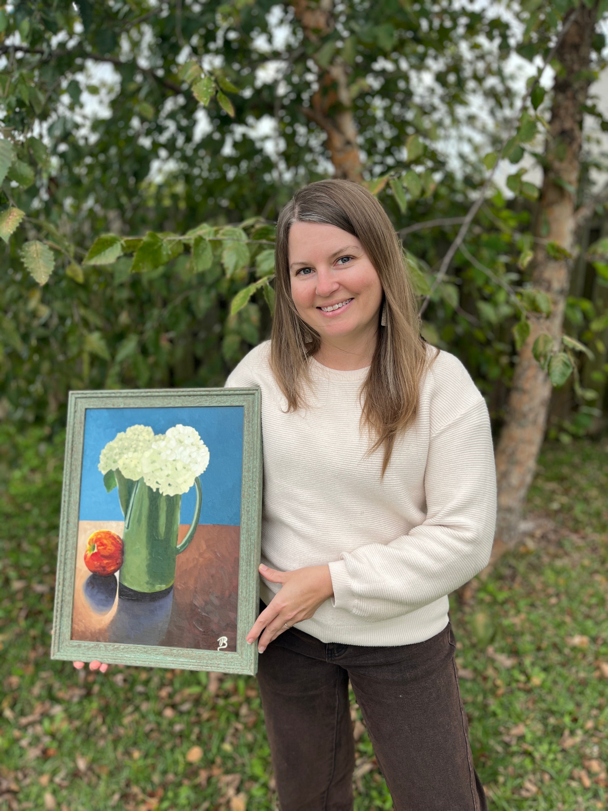 Artist, Blair Owens, holding a framed still life painting of hydrangea flowers and an apple outdoors.