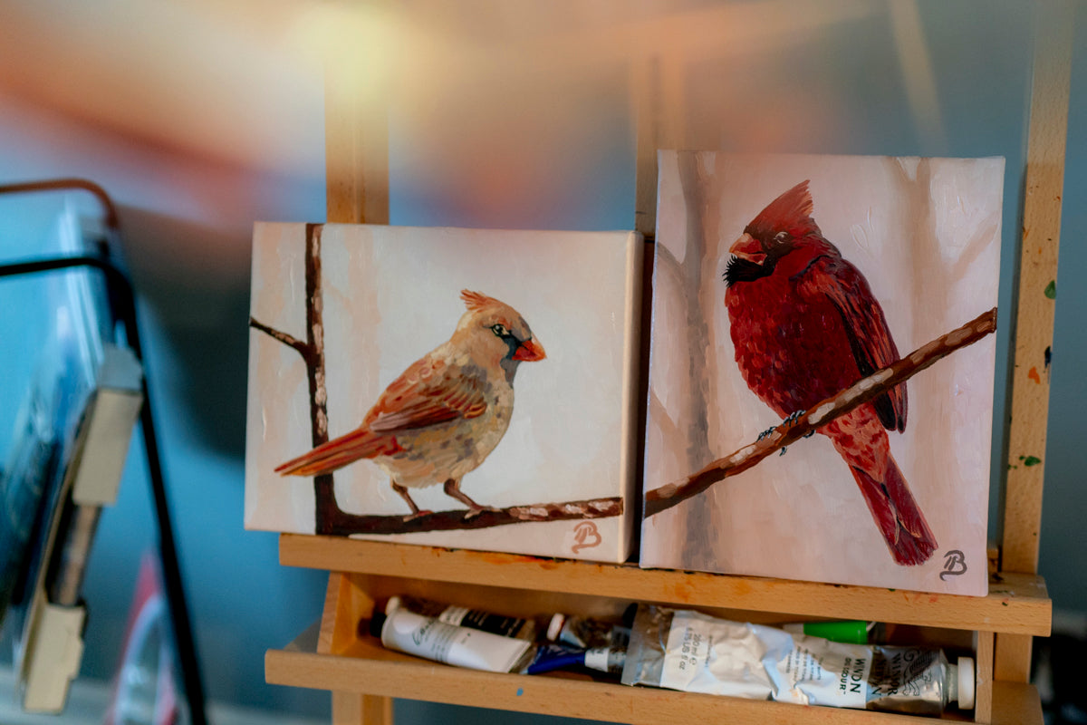Two paintings of cardinal birds on a wooden easel with art supplies.