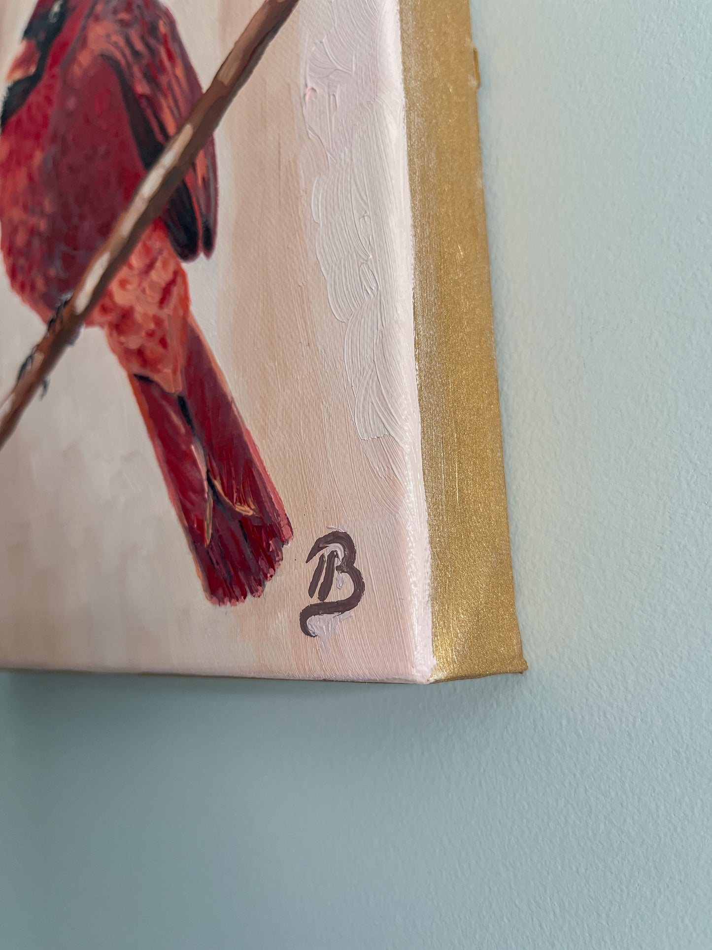 Close-up of a painting of a red cardinal bird on a branch with a gold painted canvas edge against a light gray wall.