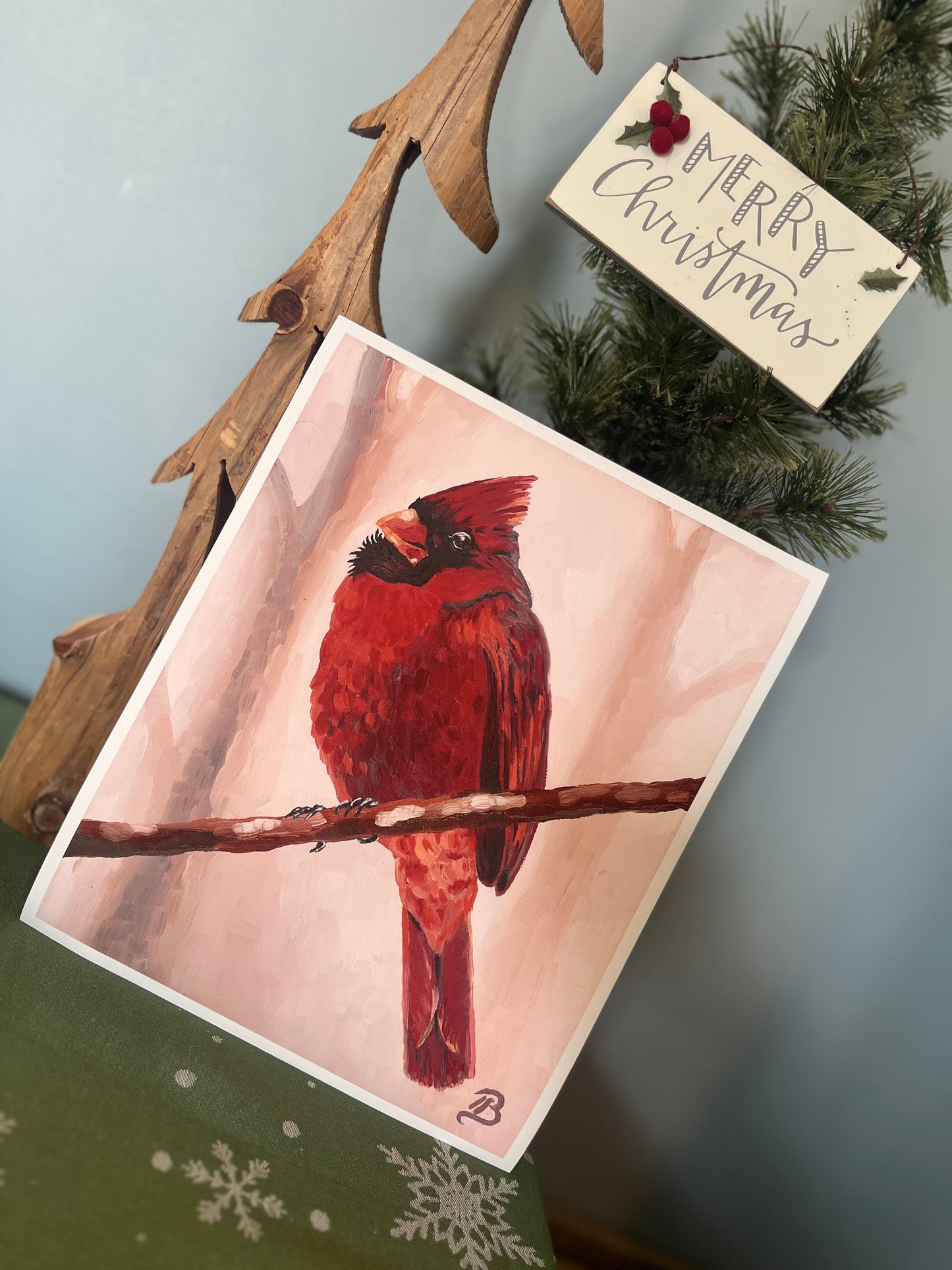Fine art print of a red cardinal perched on a branch with a soft pink background propped against Christmas tree decor