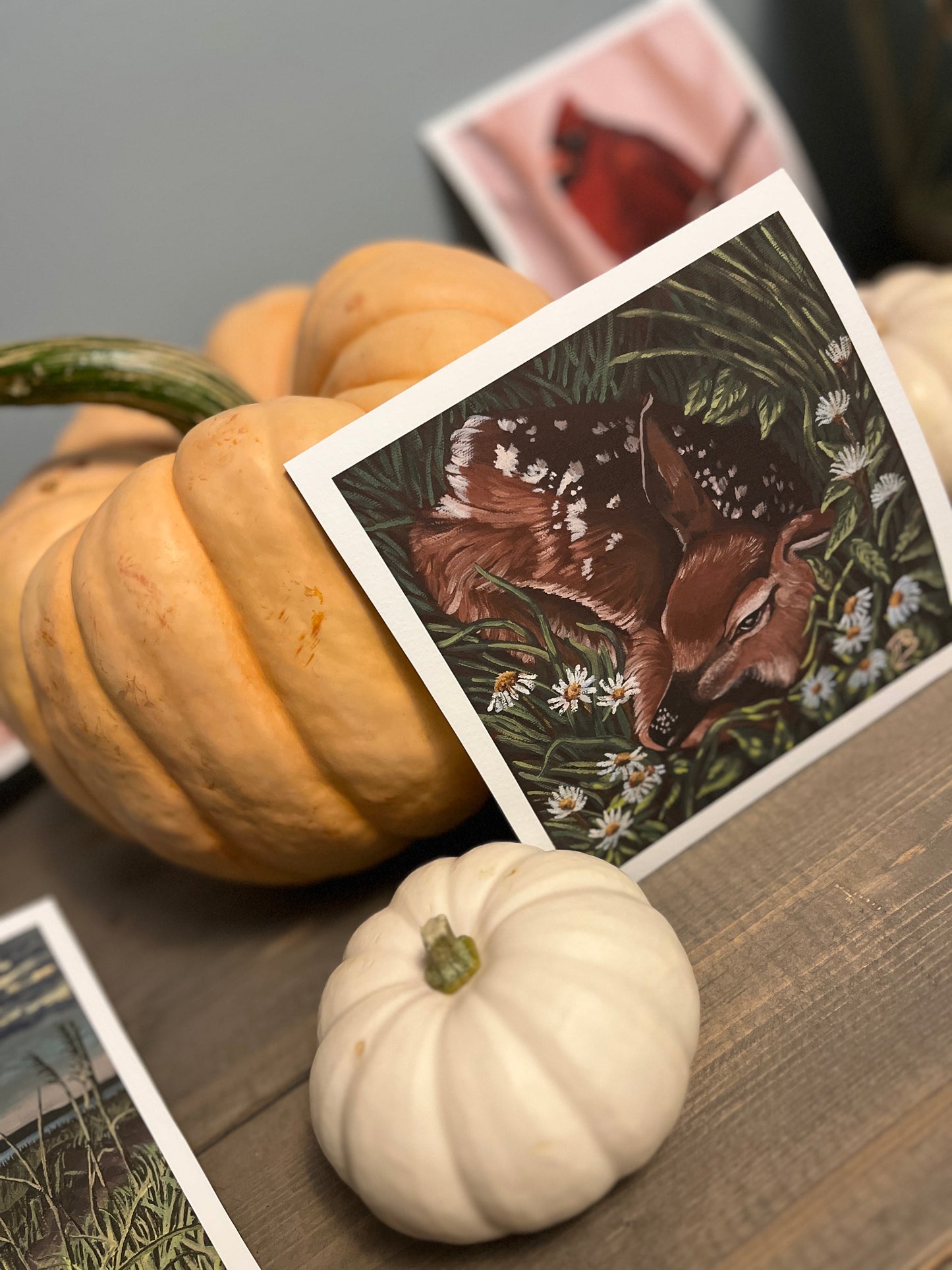 Fine art print on watercolor paper of a deer nestled in a meadow of flowers. Pumpkins are decorating the table where the art print is propped.