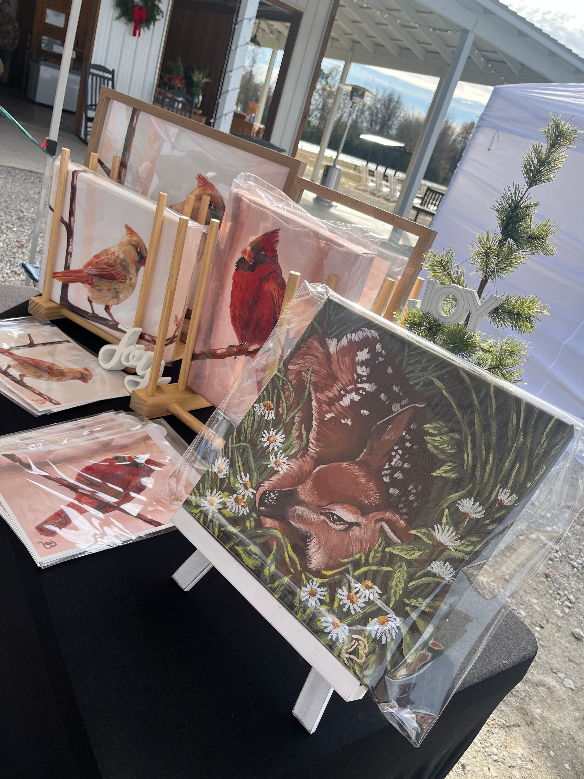 Collection of nature-themed paintings of cardinals and deer on easels outdoors.
