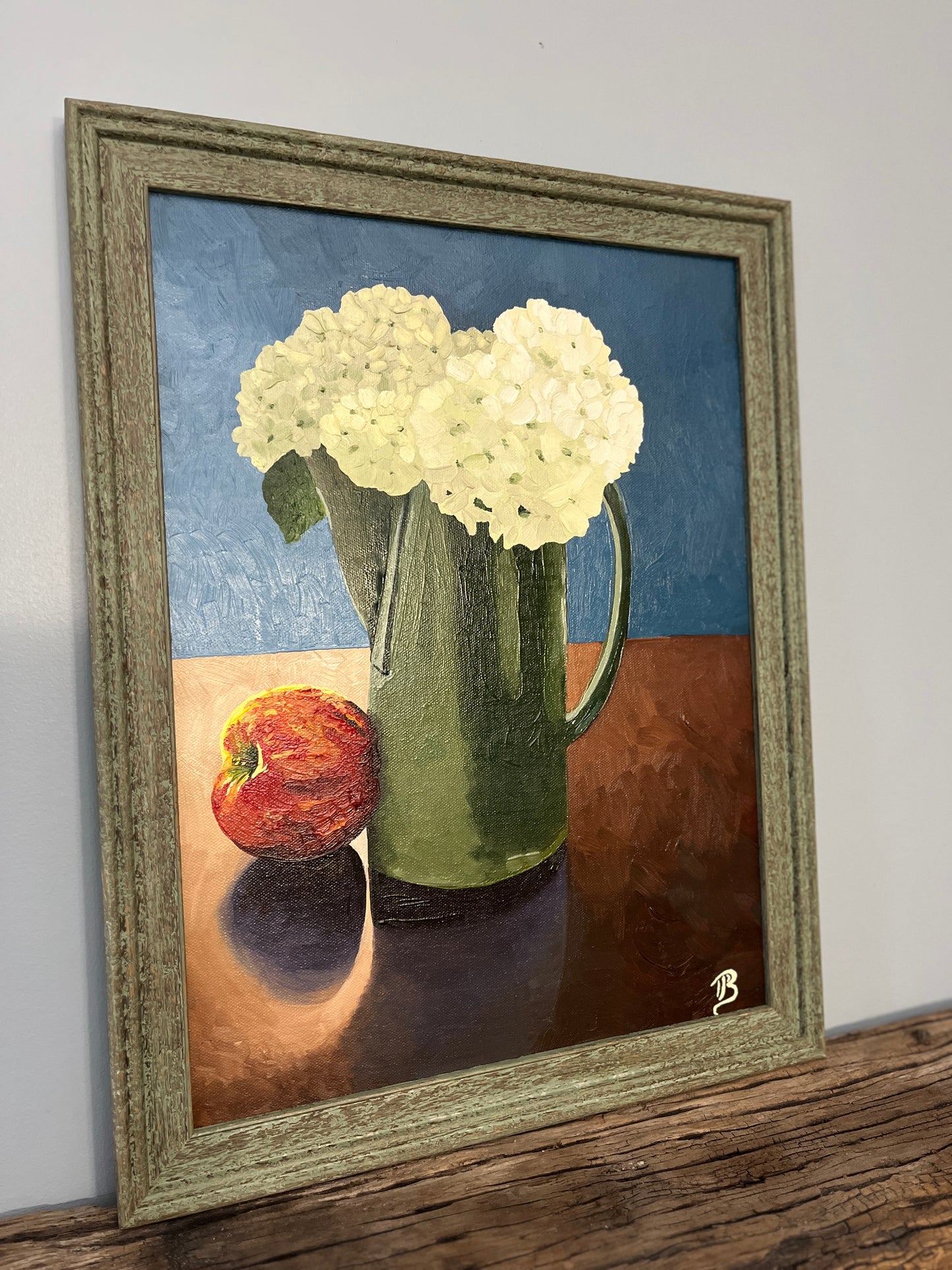 Framed still life painting with hydrangea flowers in a green pitcher and an apple on a wooden surface.