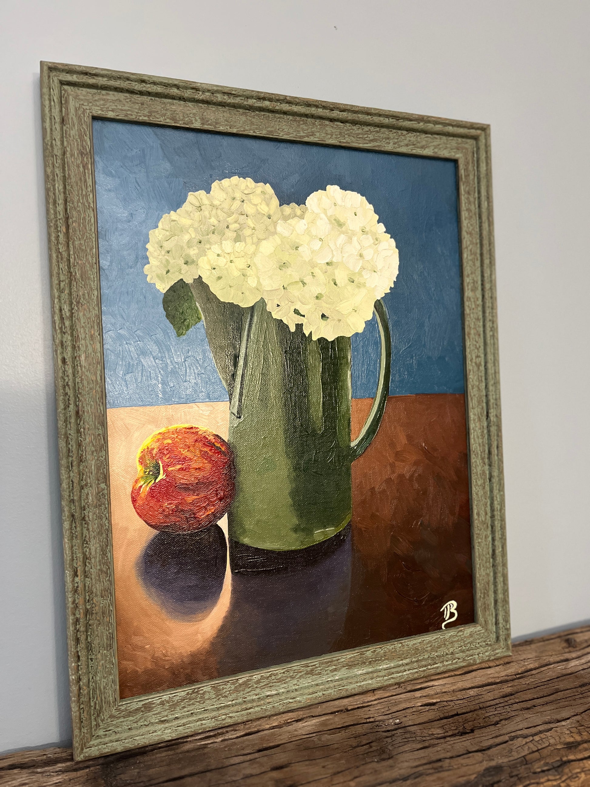 Framed still life painting with hydrangea flowers in a green pitcher and an apple on a wooden surface.