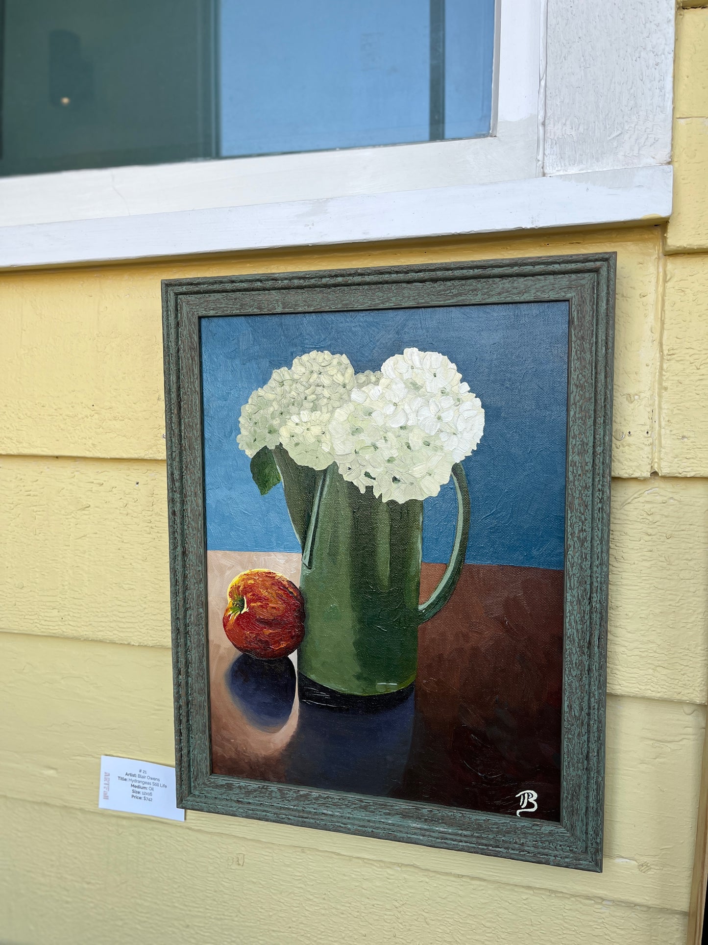 Framed painting of a green pitcher with white hydrangea flowers and an apple on a blue background.