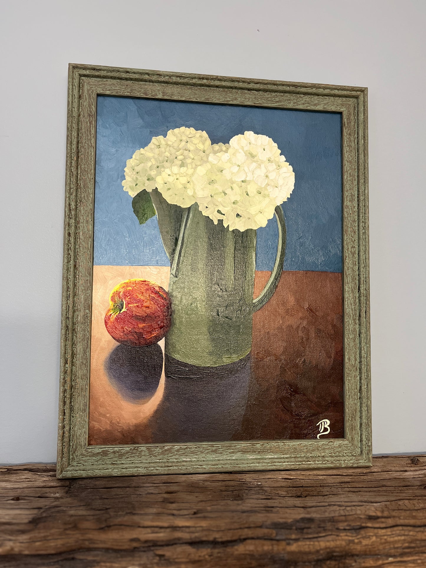 Framed artwork of a still life with hydrangea flowers in a green pitcher and an apple on a wooden surface.