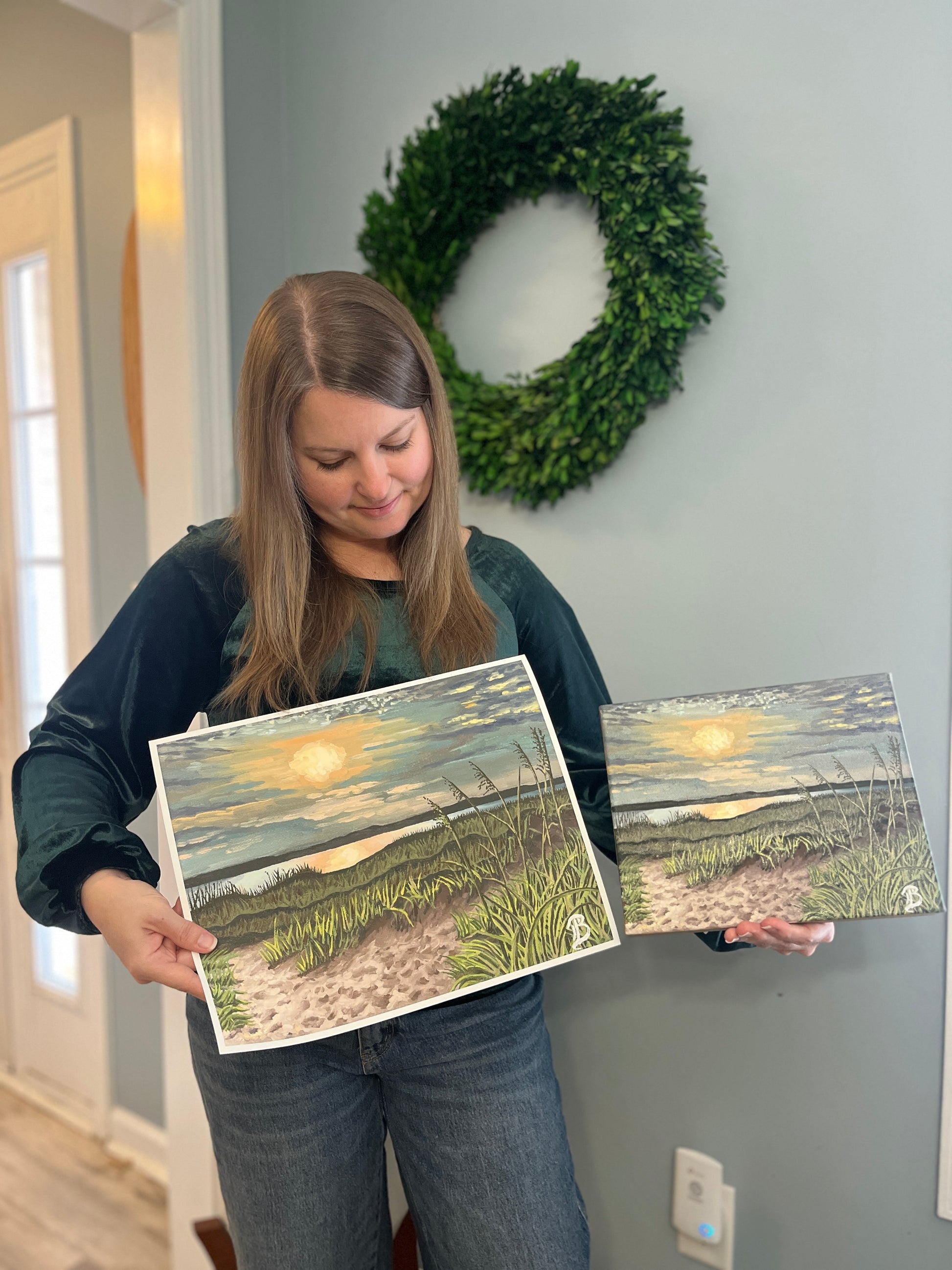 Woman holding an original painting in one hand and its fine art print in the other hand. The artwork is of a seascape sunset scene with a wreath on the wall behind her.