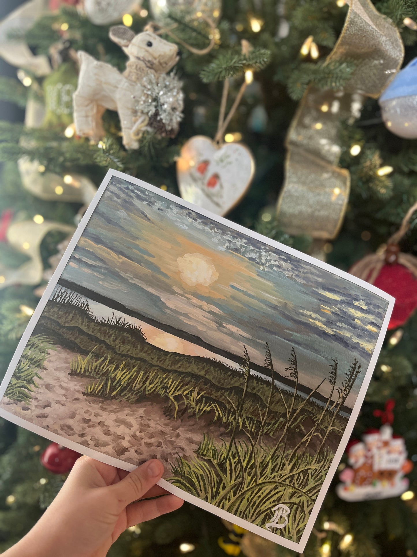 Hand holding a fine art print of a sunset beach scene in front of a decorated Christmas tree.