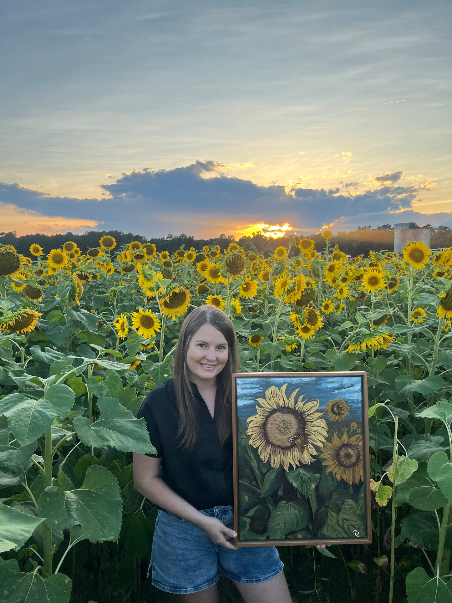Artist, Blair Owens, holding her original oil painting of a sunflower field while standing in a field of sunflowers at sunset.