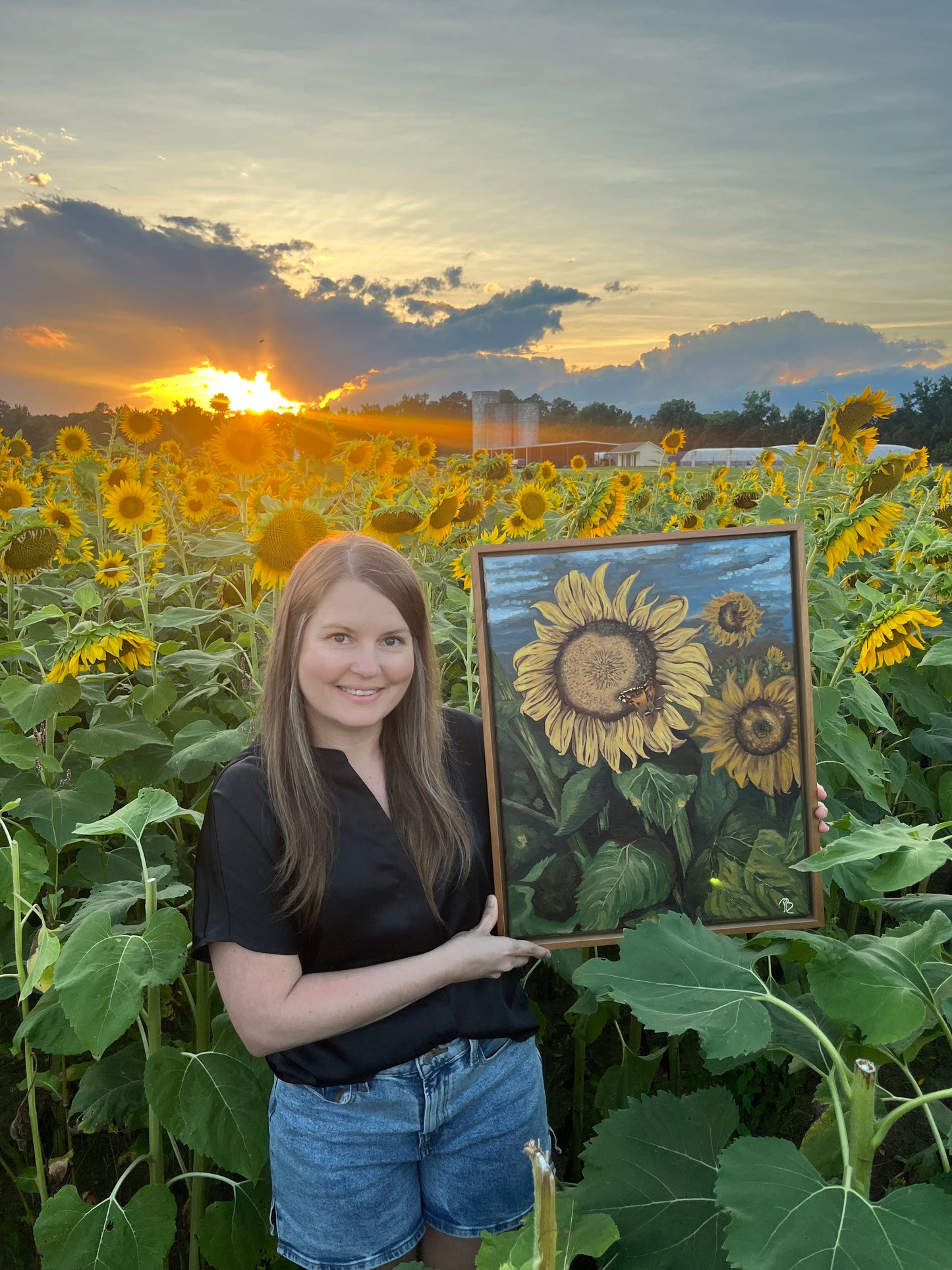 Artist, Blair Owens, holding her original oil painting of a sunflower field while standing in a field of sunflowers at sunset.