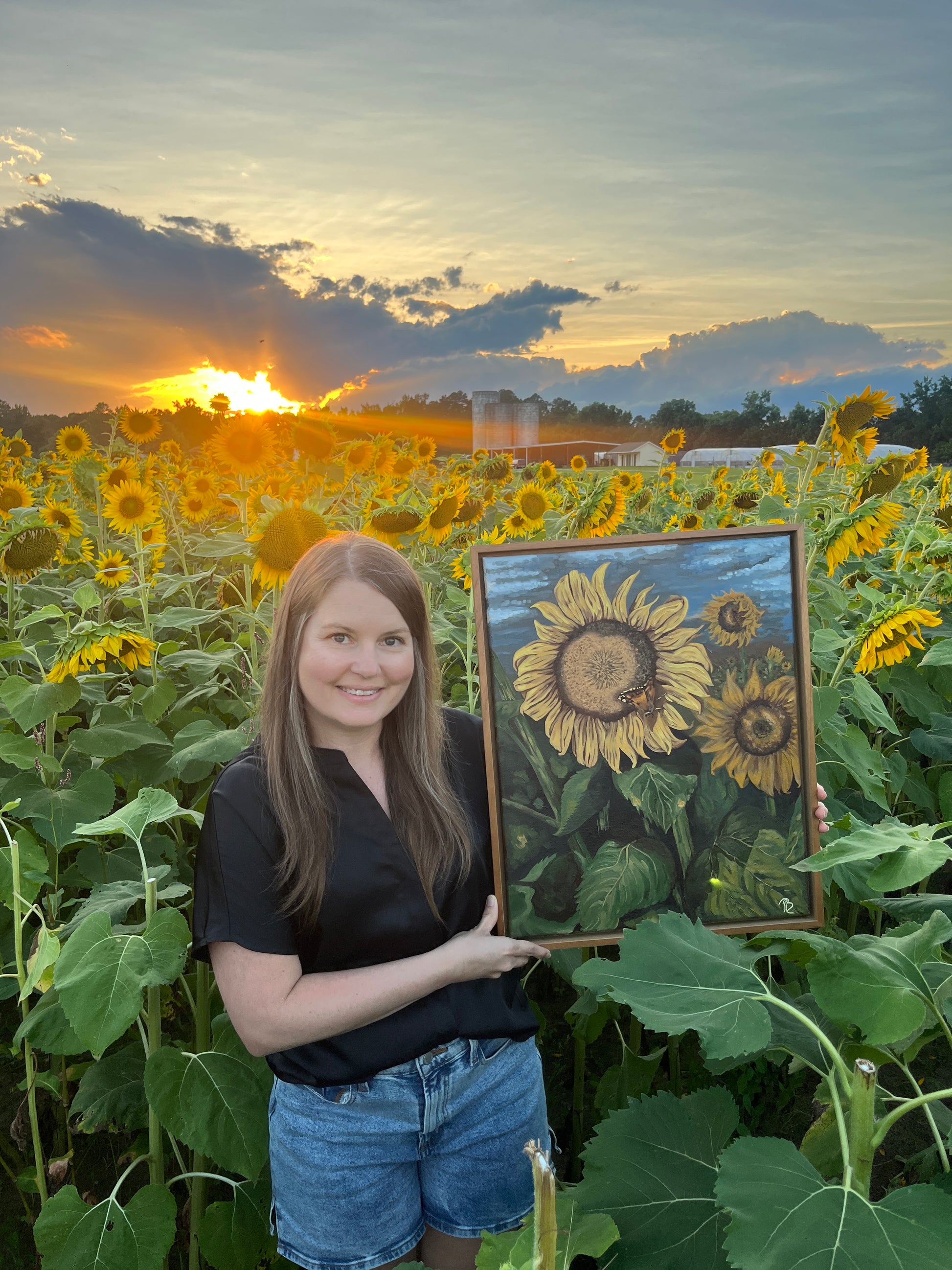 Artist, Blair Owens, holding her original oil painting of a sunflower field while standing in a field of sunflowers at sunset.