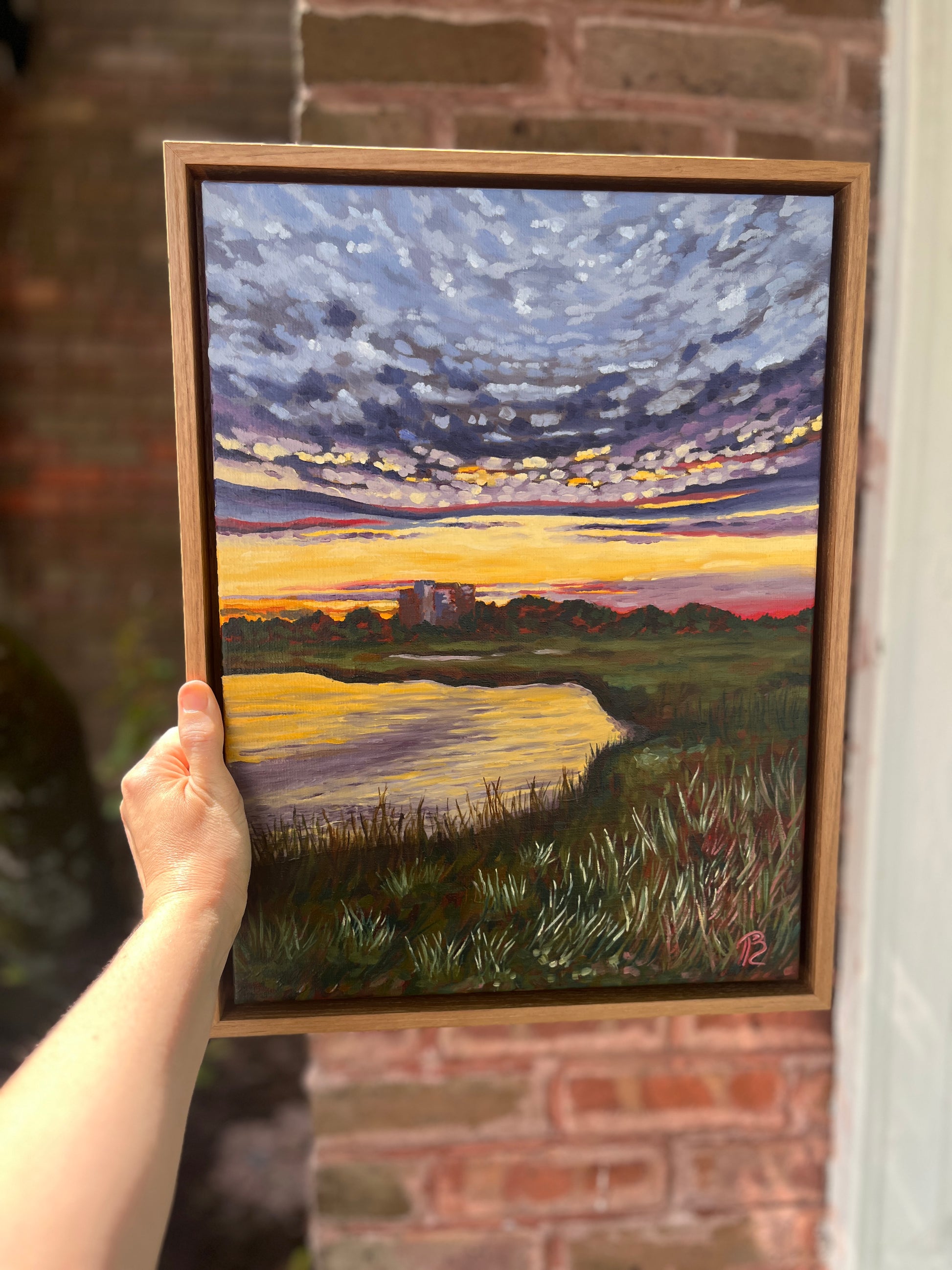 An artist's outstretched arm holding her framed painting of a sunset over the intracoastal waterway and marsh with building in distance. There is a brick wall in the background behind the painting.