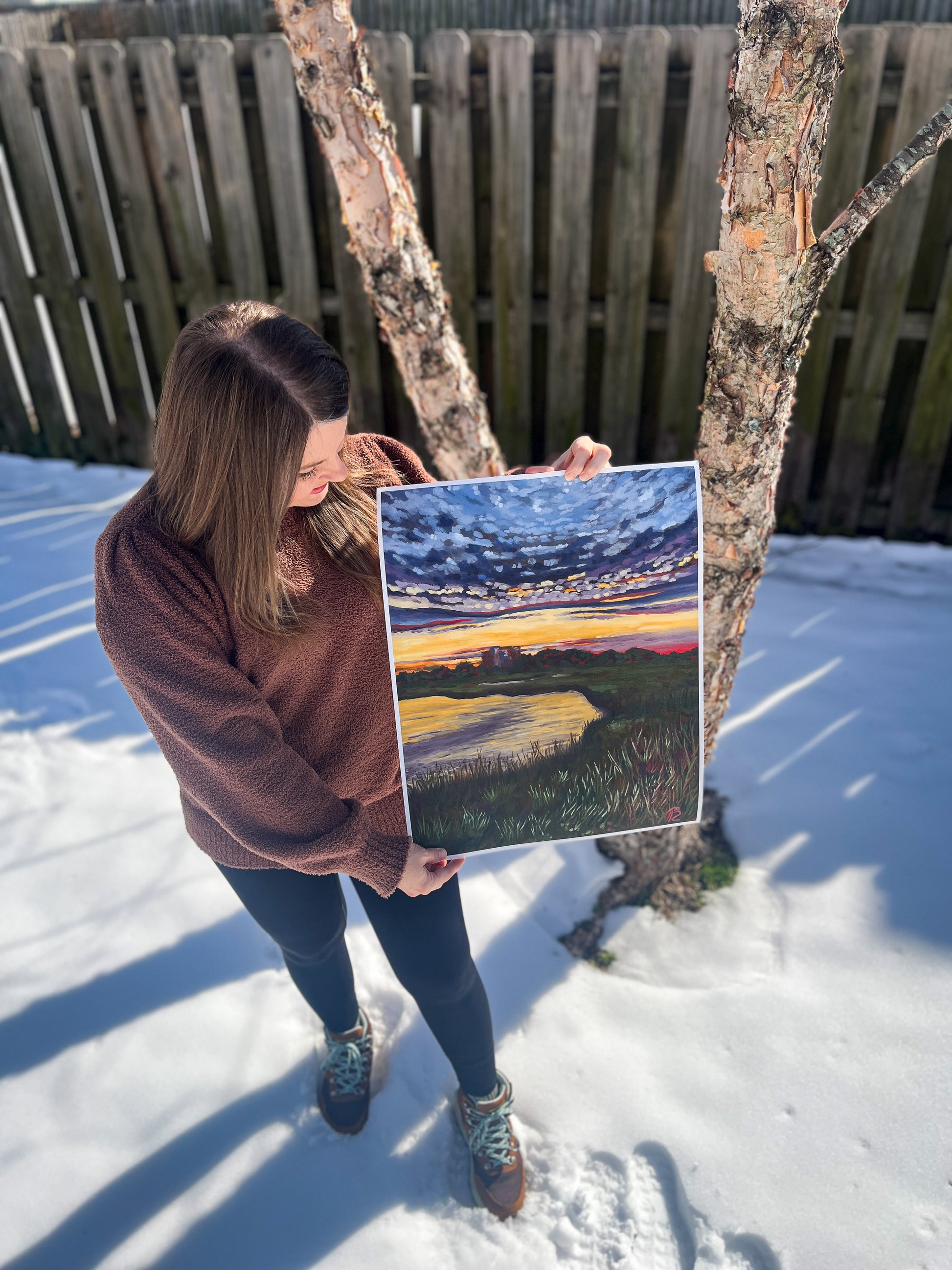 Artist holding a painting of a sunset over the marsh in a snowy outdoor setting.