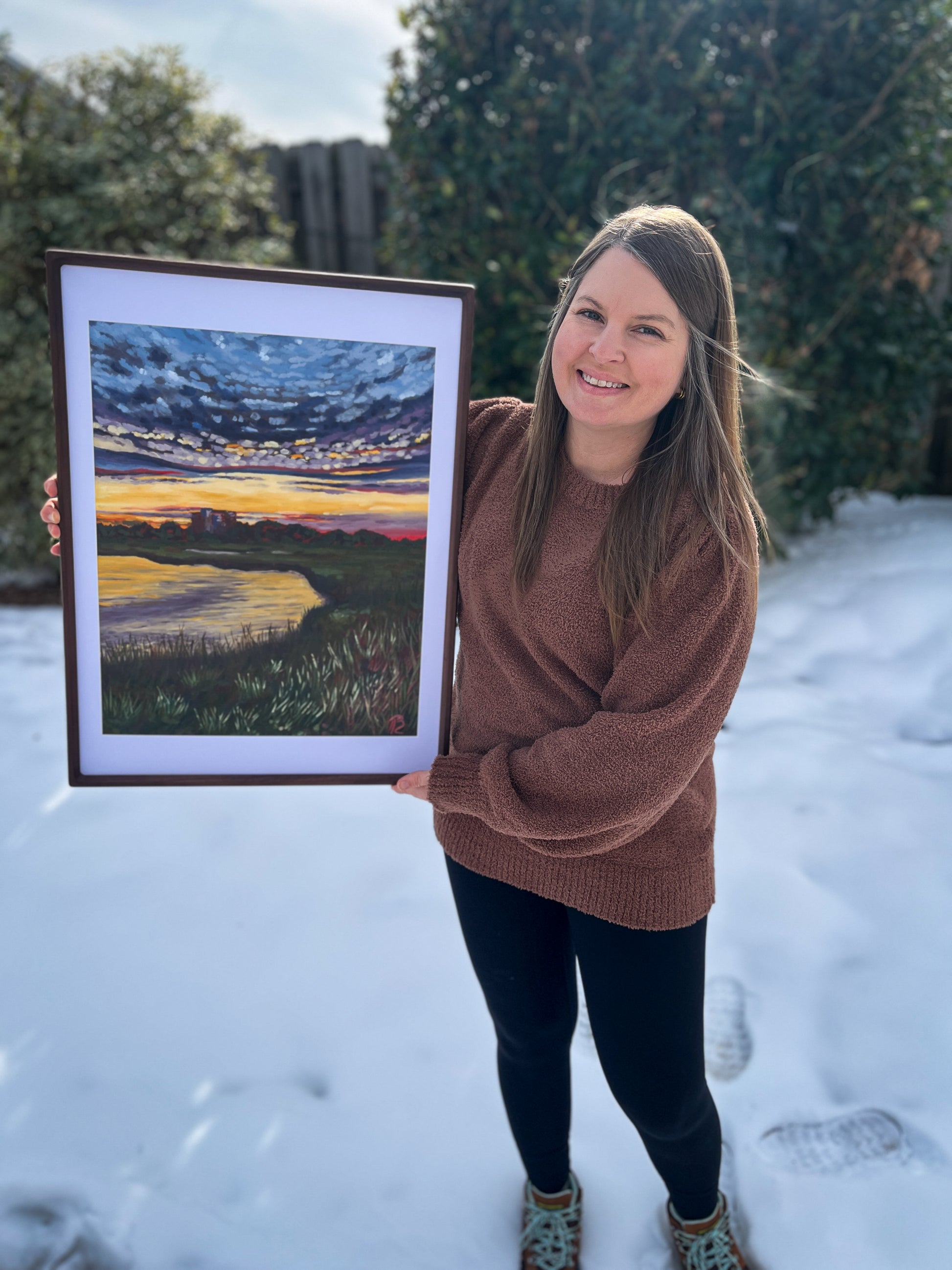 Artist, Blair Owens, holding a framed painting of sunset over the marsh while standing outdoors in the snow