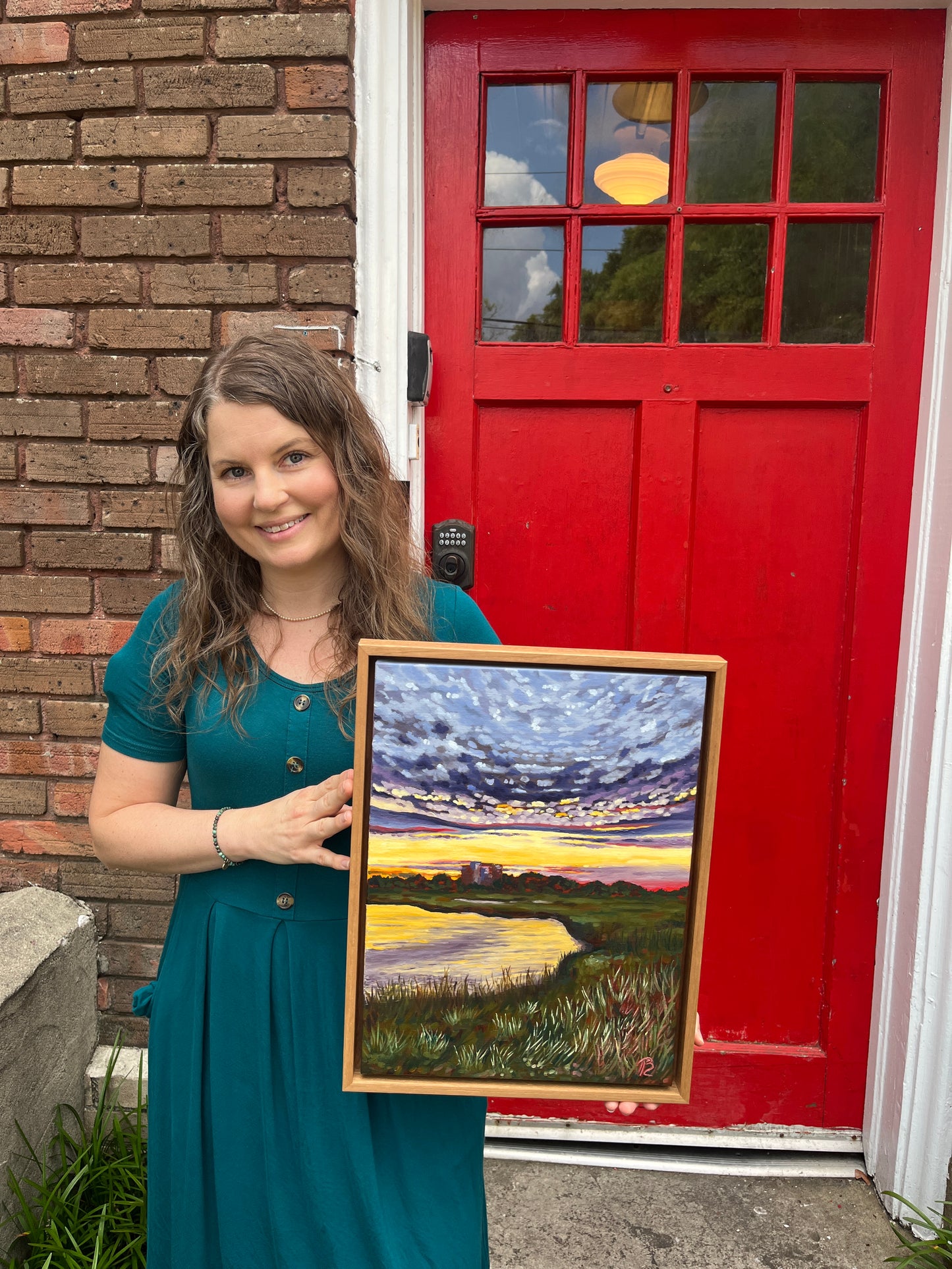 Artist, Blair Owens, holding her framed painting of a sunset over the intracoastal waterway and marsh with building in distance.